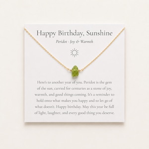 May include: A gold chain necklace featuring a small, raw green peridot gemstone pendant. The necklace is displayed on a white card printed with the text Happy Birthday, Sunshine, Peridot, Joy & Warmth, and a paragraph about the gemstone's meaning.
