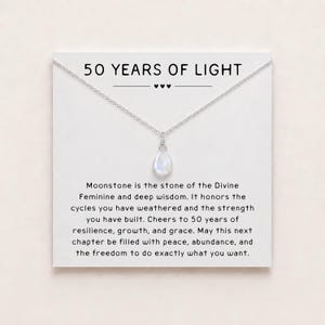 May include: A silver-toned necklace with a teardrop-shaped moonstone pendant displayed on a white card. The card features the text 50 YEARS OF LIGHT and a paragraph describing the stone, celebrating resilience, growth, and grace for a milestone.