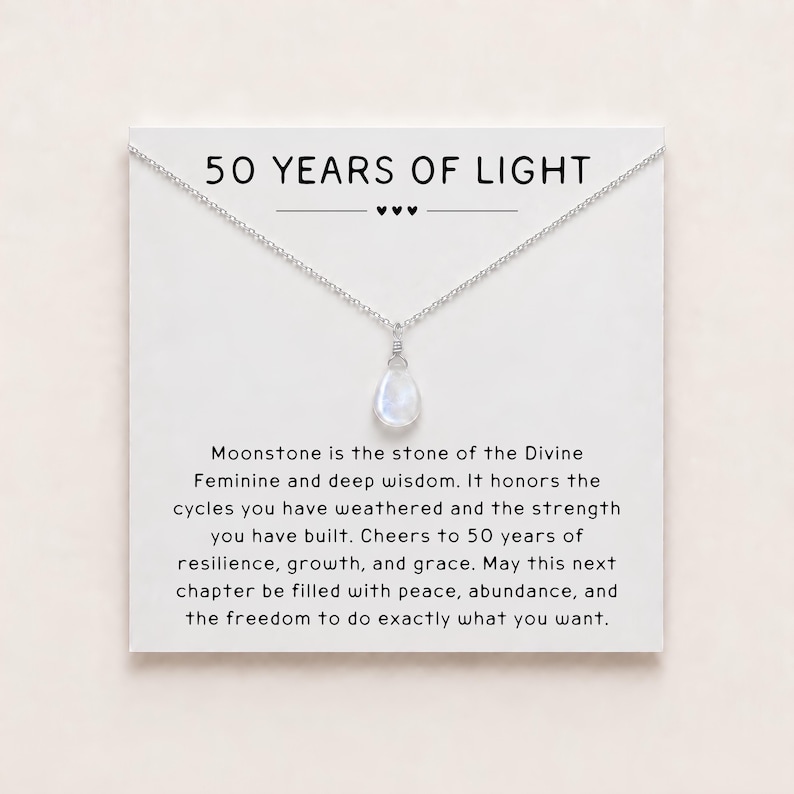 May include: A silver-toned necklace with a teardrop-shaped moonstone pendant displayed on a white card. The card features the text 50 YEARS OF LIGHT and a paragraph describing the stone, celebrating resilience, growth, and grace for a milestone.