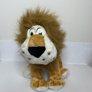 May include: A plush lion toy with a brown mane and body, white face, and large, expressive eyes. The lion has a black nose and brown spots on its face. The toy is designed to sit upright.