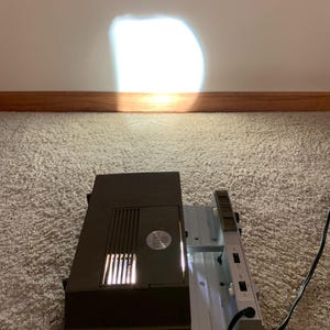 May include: A vintage brown slide projector with a bright light shining on a white wall. The projector is on a light-colored carpet. The projector has the text "Viewlex 400" on the side.