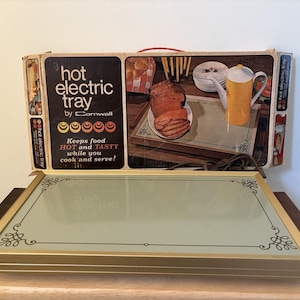 Può includere: Vassoio elettrico "Hot Electric Tray by Cornwall" vintage. Lo scaldavivande ha una cornice in metallo color oro con accenti decorativi agli angoli e manici in legno. La scatola originale è visibile, con illustrazioni di cibo e il testo "Keeps food HOT and TASTY while you cook and serve!"