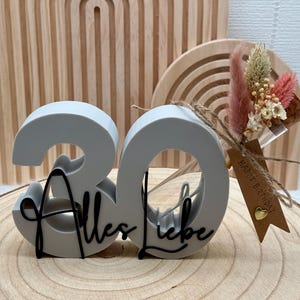 Könnte beinhalten: Eine hellgraue 3D-Zahl 30 als Dekoration mit dem schwarzen, kursiven Schriftzug Alles Liebe auf der Vorderseite. Ein kleiner Strauß aus getrockneten rosa und weißen Blumen ist mit einer Schnur befestigt, inklusive eines braunen Anhängers mit Happy Birthday.
