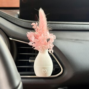 Könnte beinhalten: Ein kleiner weißer Auto-Lüftungsschlitz-Clip mit geripptem Vasendesign und Herz-Symbol, bestückt mit einem Strauß getrockneter rosa Blumen, darunter Hasenschwanzgras und Hortensienblüten, befestigt an einer horizontalen schwarzen Lüftungsdüse.