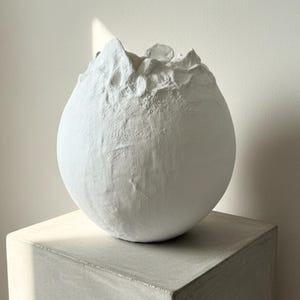 Puede incluir: Un jarrón esférico blanco texturizado con una parte superior rugosa e irregular. El jarrón se encuentra sobre un pedestal cuadrado de color gris claro. La iluminación resalta la textura del jarrón y la forma geométrica del pedestal.