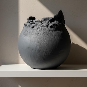 Op de afbeelding: Een donkergrijze, bolvormige keramische vaas met een getextureerde, organische rand. De vaas staat op een witte plank tegen een neutraal getinte muur. De ruwe textuur van de rand contrasteert met de gladde, afgeronde body. De vaas heeft een diameter van ongeveer 25 cm.