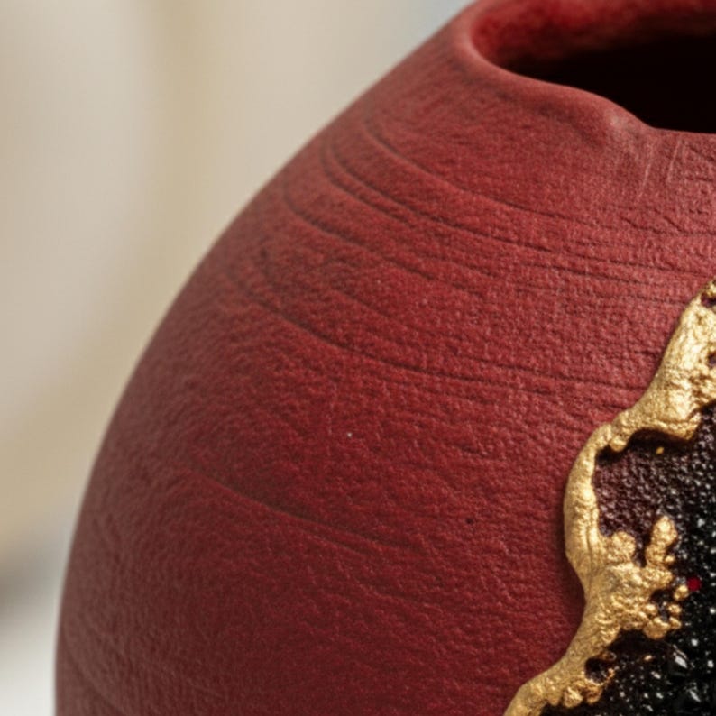 May include: Close-up of a textured, maroon-colored ceramic vase. The vase has a rounded shape with a slightly flared opening. A gold-colored, ornate design adorns a portion of the vase, adding a decorative element.