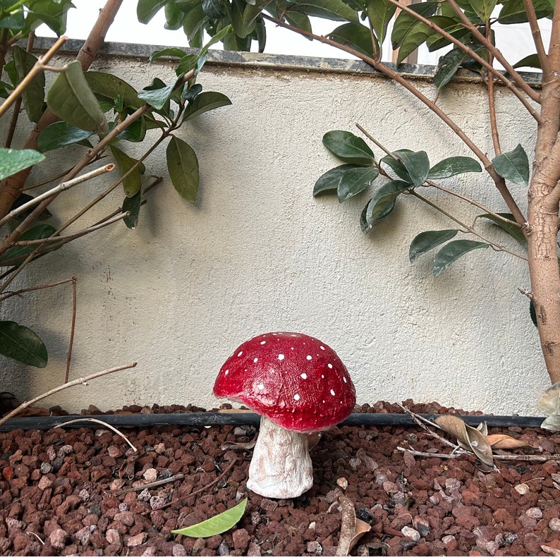 May include: A red and white mushroom shaped garden statue with white polka dots. The statue is sitting on a bed of small brown rocks.