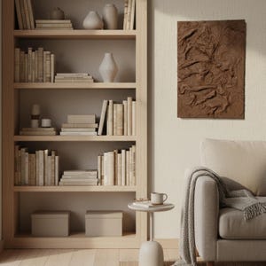 May include: A wooden bookcase filled with books and vases. A brown textured artwork hangs on the wall. A small side table with a mug and a sofa with a blanket complete the room.