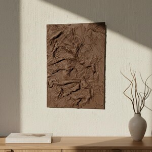 May include: A rectangular, brown textured wall art piece with a relief design, mounted on a cream-coloured wall. The artwork is the focal point, with a neutral-toned vase and a book on a wooden surface below. Natural light casts shadows.