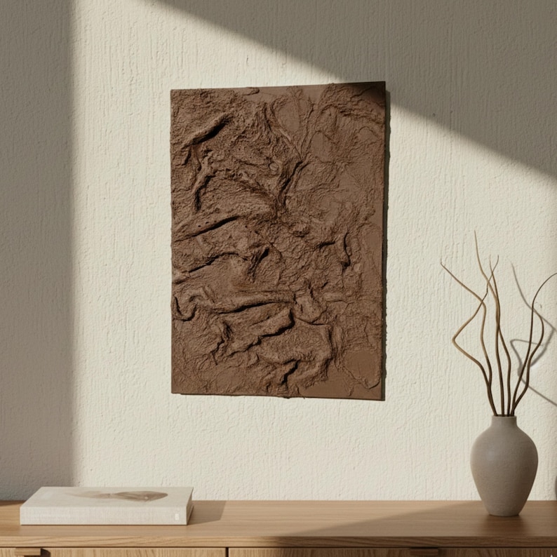 May include: A rectangular, brown textured wall art piece with a relief design, mounted on a cream-coloured wall. The artwork is the focal point, with a neutral-toned vase and a book on a wooden surface below. Natural light casts shadows.