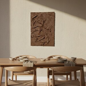 May include: A textured brown rectangular artwork is mounted on a wall above a wooden dining table. The table is set with plates, bowls, and napkins. Wooden chairs are arranged around the table.
