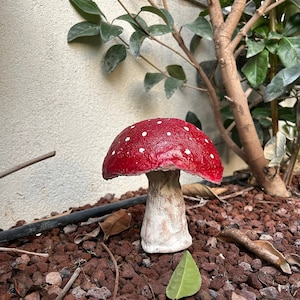 May include: A red and white mushroom figurine with white dots on the top. The mushroom is sitting on a bed of red gravel.