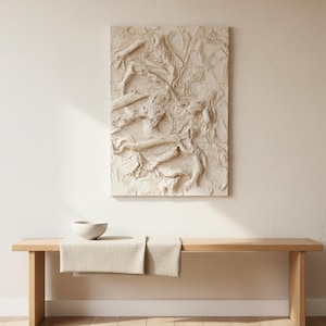 May include: An off-white, textured abstract artwork hangs above a wooden bench. The rectangular piece has a raised design. A small, light-coloured bowl sits on the bench, with a beige runner draped across it. The scene is lit by natural light.
