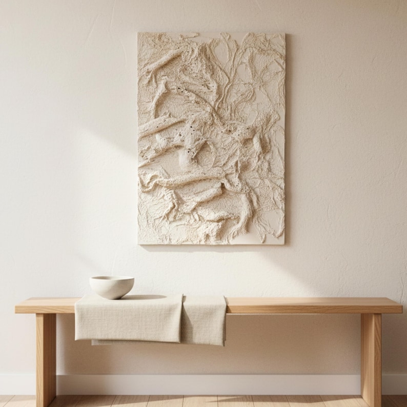 May include: An off-white, textured abstract artwork hangs above a wooden bench. The rectangular piece has a raised design. A small, light-coloured bowl sits on the bench, with a beige runner draped across it. The scene is lit by natural light.