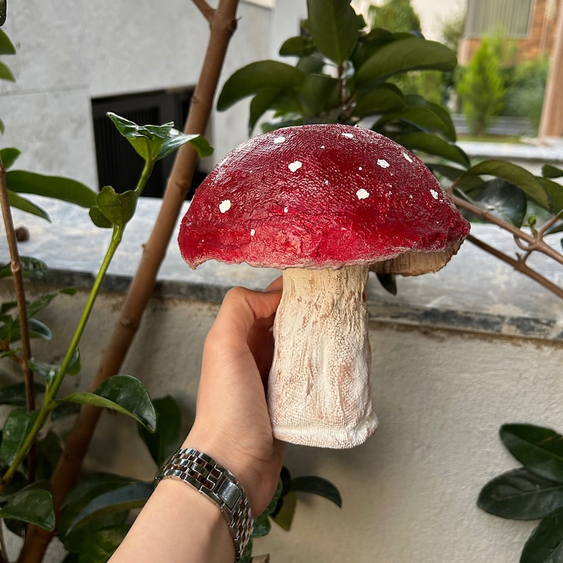 May include: A red and white mushroom figurine with white spots on the cap. The mushroom is made of a textured material and has a white stem.
