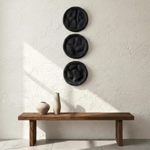 May include: Three black, textured, circular wall art pieces are mounted on a white wall. Below, a wooden bench holds two neutral-toned vases. The bench has a natural wood grain finish and a simple, modern design.