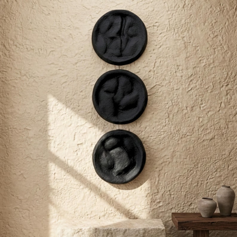 May include: Three black, circular wall art pieces with textured surfaces and abstract designs. The art is mounted on a textured, beige wall. Decorative pottery sits on a wooden table in the background.