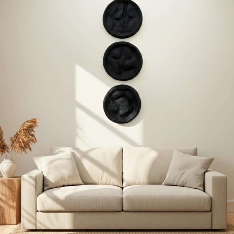 May include: Three black decorative wall plates with textured surfaces are mounted vertically on a cream-colored wall. Below, a beige sofa with matching pillows sits in front of a wooden side table with a vase of dried plants.