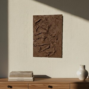 May include: A rectangular, textured brown wall art piece with a relief design. The artwork is mounted on a light beige wall. A wooden cabinet with books and a vase is in the foreground.