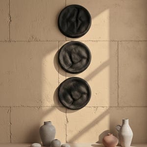 May include: Three black, textured, circular wall decorations are mounted on a beige wall. Below, a collection of decorative vases and small stones in neutral tones are arranged on a wooden surface. The scene is bathed in natural light.