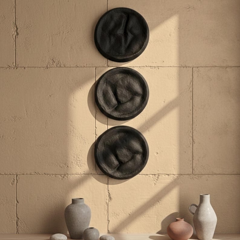 May include: Three black, textured, circular wall decorations are mounted on a beige wall. Below, a collection of decorative vases and small stones in neutral tones are arranged on a wooden surface. The scene is bathed in natural light.