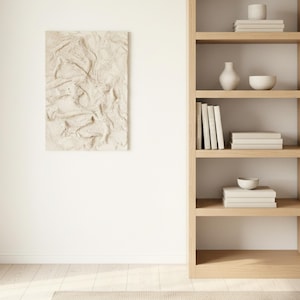 May include: A textured, neutral-toned artwork is mounted on a white wall. A wooden bookshelf on the right holds books, a vase, and bowls. The room has a minimalist, modern design with light wood and neutral colours.