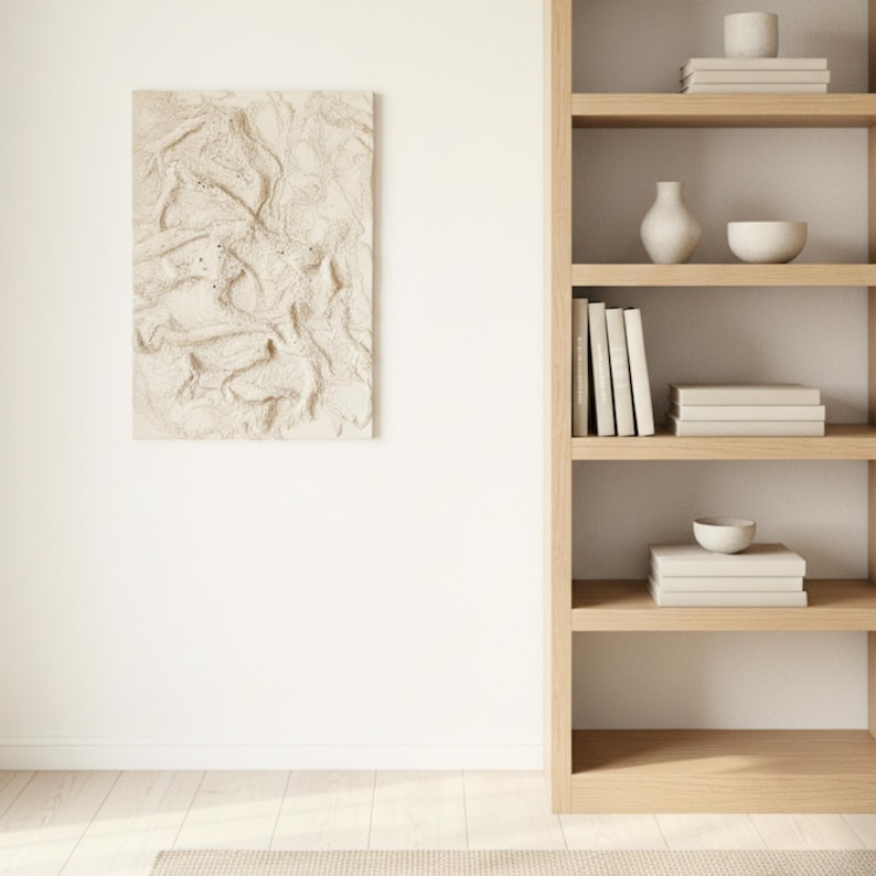 May include: A textured, neutral-toned artwork is mounted on a white wall. A wooden bookshelf on the right holds books, a vase, and bowls. The room has a minimalist, modern design with light wood and neutral colours.