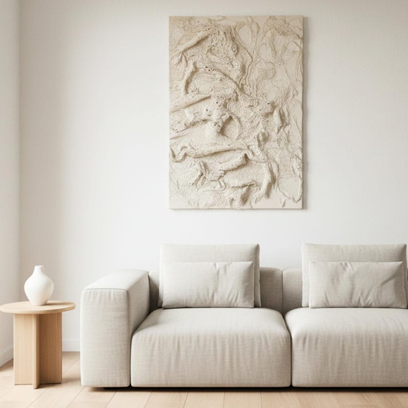 May include: A modern living room with a beige sofa and cushions, a small wooden side table, and a white vase. Above the sofa hangs a large, textured, beige wall art piece, adding a contemporary touch to the room.