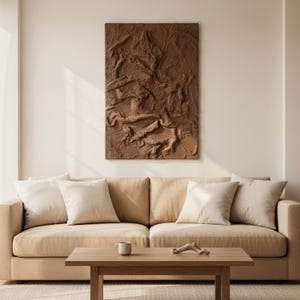 May include: A brown textured wall art piece hangs above a beige sofa with several white cushions. A wooden coffee table in front of the sofa holds a small white mug and a decorative wooden piece. The room has a neutral colour palette.