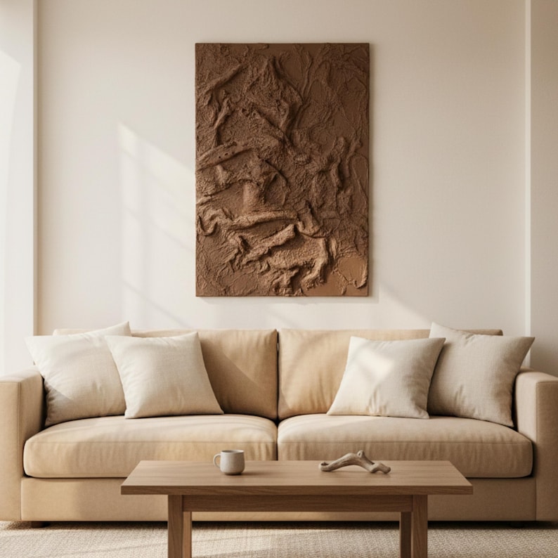 May include: A brown textured wall art piece hangs above a beige sofa with several white cushions. A wooden coffee table in front of the sofa holds a small white mug and a decorative wooden piece. The room has a neutral colour palette.