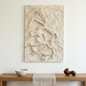 May include: An off-white textured abstract artwork hangs above a wooden table. A small, light-coloured bowl and a dark wooden sculpture sit on the table. A beige runner is placed on the table.