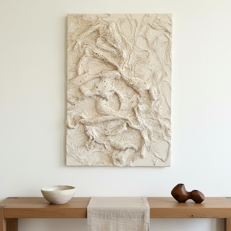 May include: An off-white textured abstract artwork hangs above a wooden table. A small, light-coloured bowl and a dark wooden sculpture sit on the table. A beige runner is placed on the table.