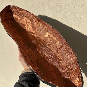 May include: A long, oval-shaped bowl made of paper mache with a copper-colored finish. The bowl has a textured surface and is slightly curved.
