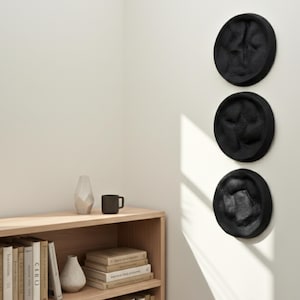 May include: Three black, circular wall sculptures with textured surfaces are mounted on a white wall. The sculptures are arranged vertically. A wooden bookshelf with books and decorative items is in the background.