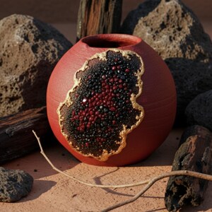 May include: A round, red ceramic vase with a unique design. A section of the vase is filled with black and red stones, outlined with gold. The vase is surrounded by rocks and wood.