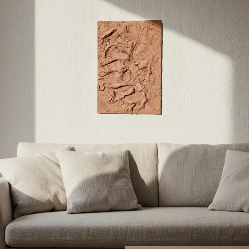 May include: A terracotta-coloured, textured rectangular wall art piece is displayed above a neutral sofa with cushions. The artwork features a raised, organic design, adding visual interest. The room is illuminated by natural light, creating a warm atmosphere.