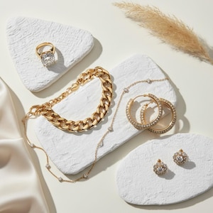 May include: An assortment of gold-toned jewelry, including a ring with a large square stone, a chunky chain bracelet, a delicate necklace, hoop earrings, and stud earrings. The jewelry is displayed on white, textured platforms.