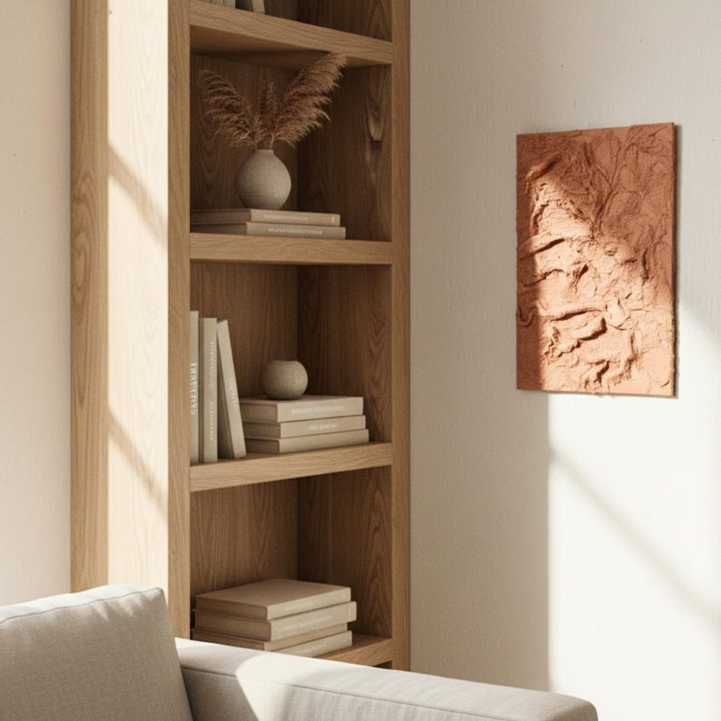 May include: A light-coloured wooden bookshelf with multiple shelves. The shelves display books, decorative vases, and stacks of books. A textured, reddish-brown wall art piece hangs on the wall to the right.
