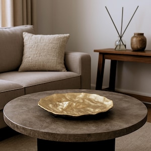 Könnte beinhalten: Eine goldfarbene Dekoschale mit strukturierter Oberfläche steht auf einem dunkelgrauen runden Couchtisch. Die Schale hat eine organische, wellenförmige Form. Im Hintergrund sind ein beiges Sofa und Kissen sowie ein Beistelltisch aus Holz und Dekorationsgegenstände zu sehen.