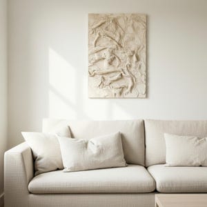 May include: A beige, textured wall art piece with an abstract design hangs above a beige sofa with matching cushions. The artwork has a raised, organic pattern, creating a tactile visual element. The room is bathed in natural light.