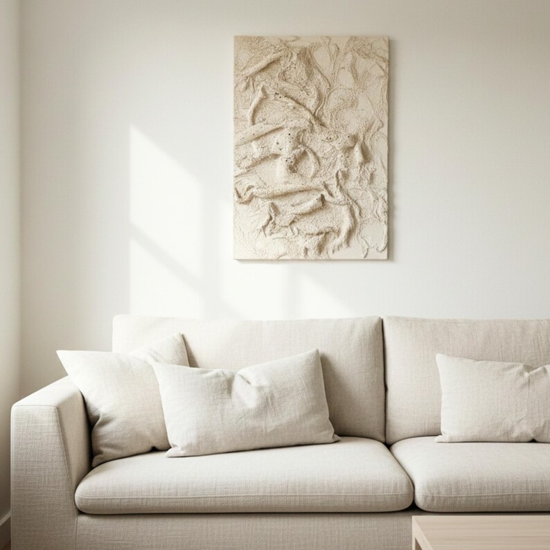 May include: A beige, textured wall art piece with an abstract design hangs above a beige sofa with matching cushions. The artwork has a raised, organic pattern, creating a tactile visual element. The room is bathed in natural light.
