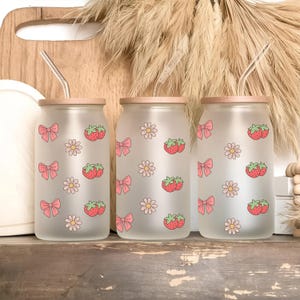 Puede incluir: Tres vasos de vidrio esmerilado con tapas de madera y pajitas de vidrio. Cada vaso está decorado con lazos rosas, fresas y diseños de margaritas. Los vasos se exhiben sobre una superficie de madera.