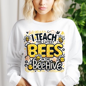 May include: White sweatshirt with a colorful graphic that reads "I teach the cutest bees in the beehive." The design features yellow honeycomb-patterned letters, cartoon bees, and floral accents. A fun and playful design for teachers.