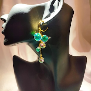 Può includere: Un paio di orecchini a cerchio dorati con una catena pendente e più perline di vetro iridescenti verdi e blu.