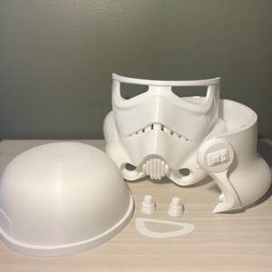 May include: A disassembled white Stormtrooper helmet, featuring the main faceplate, a separate dome, and other components. The helmet has a distinctive design with a mouth grill, eye openings, and side details. The pieces are arranged on a light-colored surface.