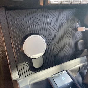 Könnte beinhalten: Ein schwarzer Auto-Konsolen-Organizer mit geometrischem Muster. Er verfügt über eine kreisförmige, silberfarbene magnetische Telefonhalterung und einen USB-Anschluss. Der Organizer ist für die Mittelkonsole eines Fahrzeugs konzipiert.