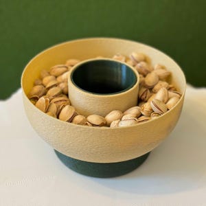 May include: A beige and green ceramic bowl with a center divider, filled with pistachios.