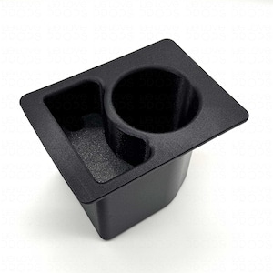 Puede incluir: Un portavasos de consola de coche de plástico negro con un portavasos circular y un compartimento más pequeño y en ángulo. El portavasos está diseñado para encajar en la consola central de un coche. El artículo está hecho de plástico negro mate.