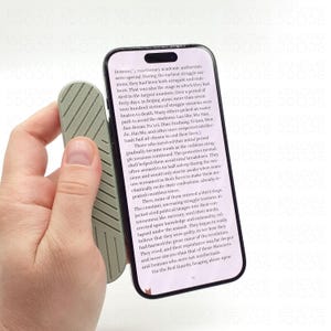 Op de afbeelding: Een smartphone vastgehouden met een lichtgroene, getextureerde telefoonhouder. De telefoon geeft tekst uit een boek weer. De houder heeft een geribbeld ontwerp. De achtergrond is wit.